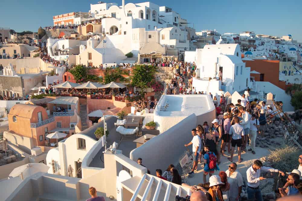 Crowded streets in Santorini, Greece, with tourists walking between whitewashed buildings and blue-domed roofs.