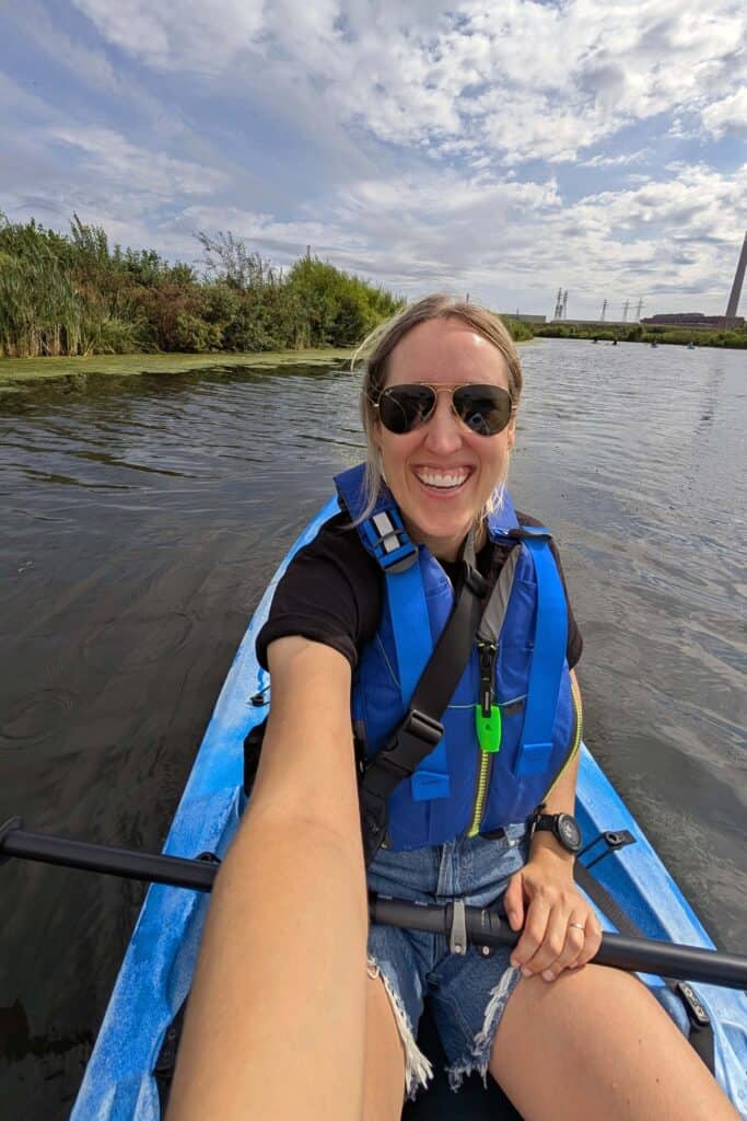 Blonde woman kayaking on the Lower Don River in Toronto, wearing a blue life jacket, with marsh vegetation and calm water surrounding the river.