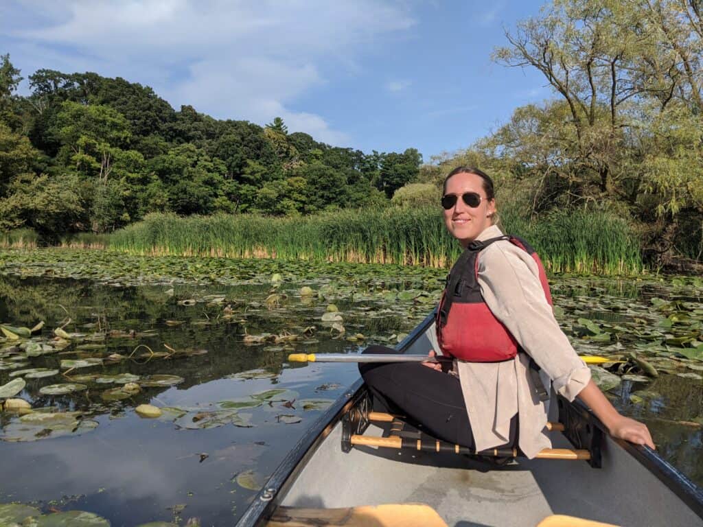 Person canoeing through a calm section of the Humber River surrounded by lily pads, cattails, and dense green forest on a sunny summer afternoon in Toronto.