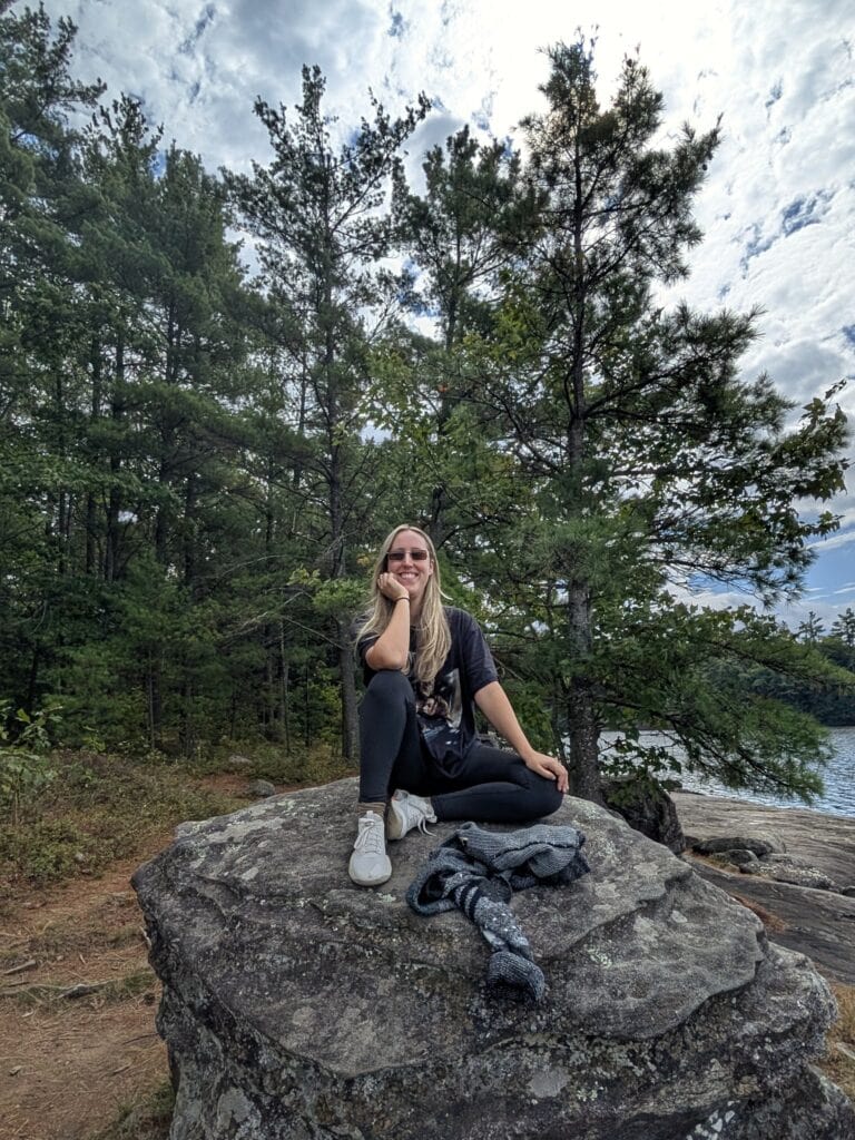 a blonde woman wearing black sitting on a large rock with trees behind her in Hardy Lake Provincial Park