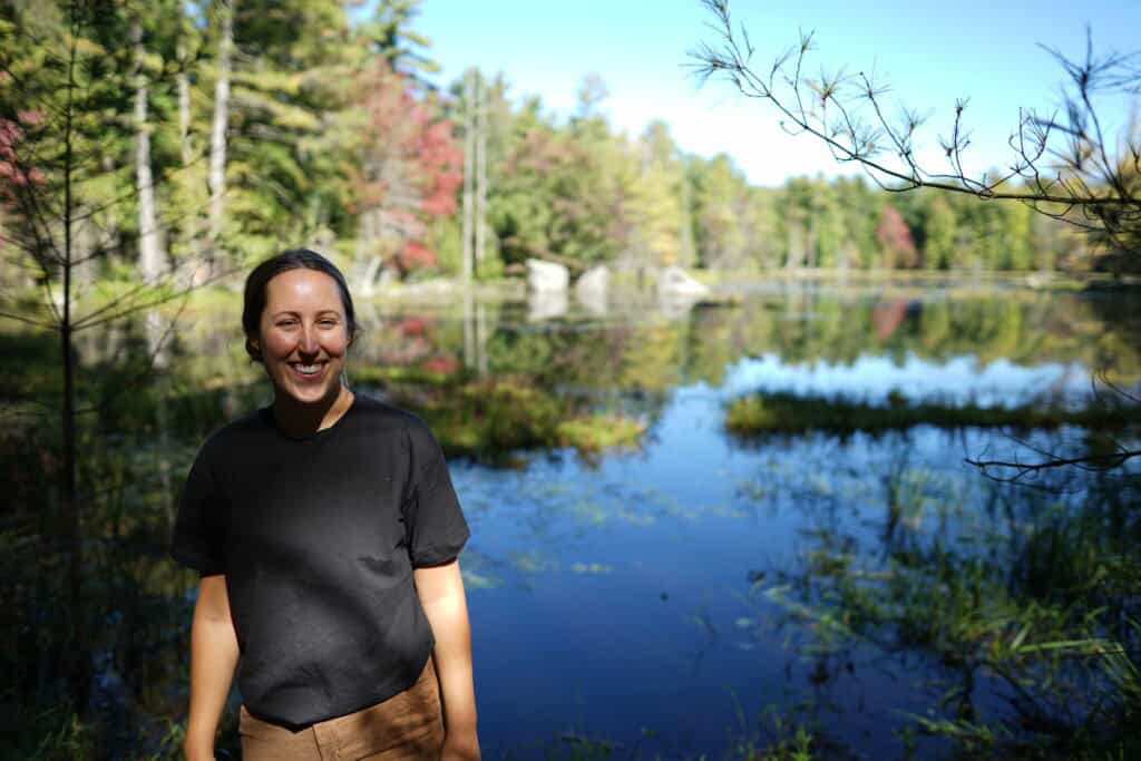 A blonde woman stands beside the calm waters of Hardy Lake in Muskoka, Ontario. The lake reflects the surrounding evergreen forest and early fall colours, while grasses and wetland vegetation line the shoreline. Large granite boulders are visible across the water beneath a clear blue sky.