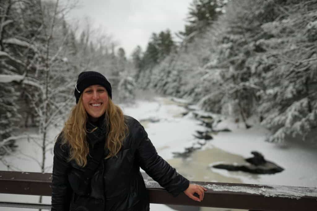 A blonde woman in a black jacket and hat stands on a bridge in Arrowhead Provincial Park during winter. Snow-covered trees line a partially frozen river below, creating a peaceful Muskoka landscape. Fresh snow blankets the forest and riverbanks beneath an overcast sky.