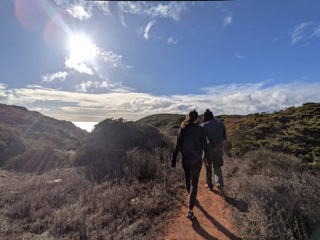 Two hikers walk along a section of Portugal's Fishermen's Trail near Sagres, following a narrow dirt path through coastal scrubland under a bright blue sky. The Atlantic Ocean is visible in the distance, with sunlight reflecting off the water.