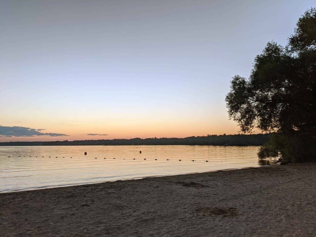 Sandy beach at Bass Lake Provincial Park at sunset. Calm water stretches across the lake as soft orange and pink hues glow along the horizon. A tree frames the shoreline, and the quiet beach sits beneath a clear evening sky.