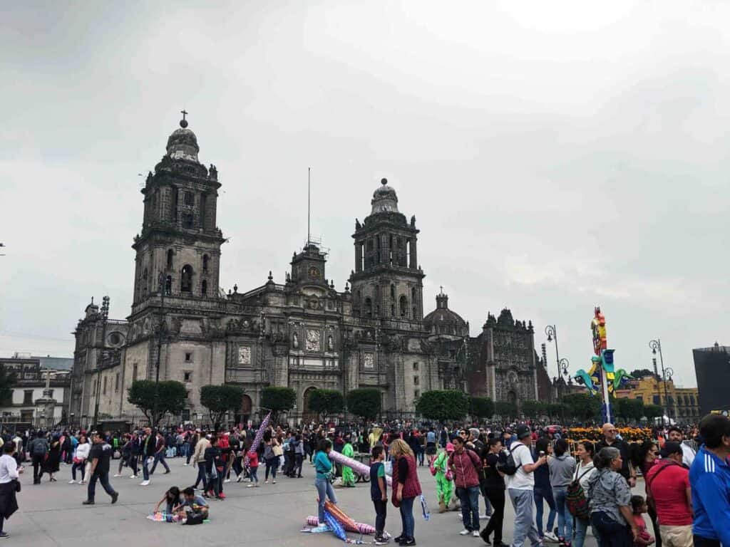 a square filled with people milling around in front of "Zocalo," a historic building in Mexico City