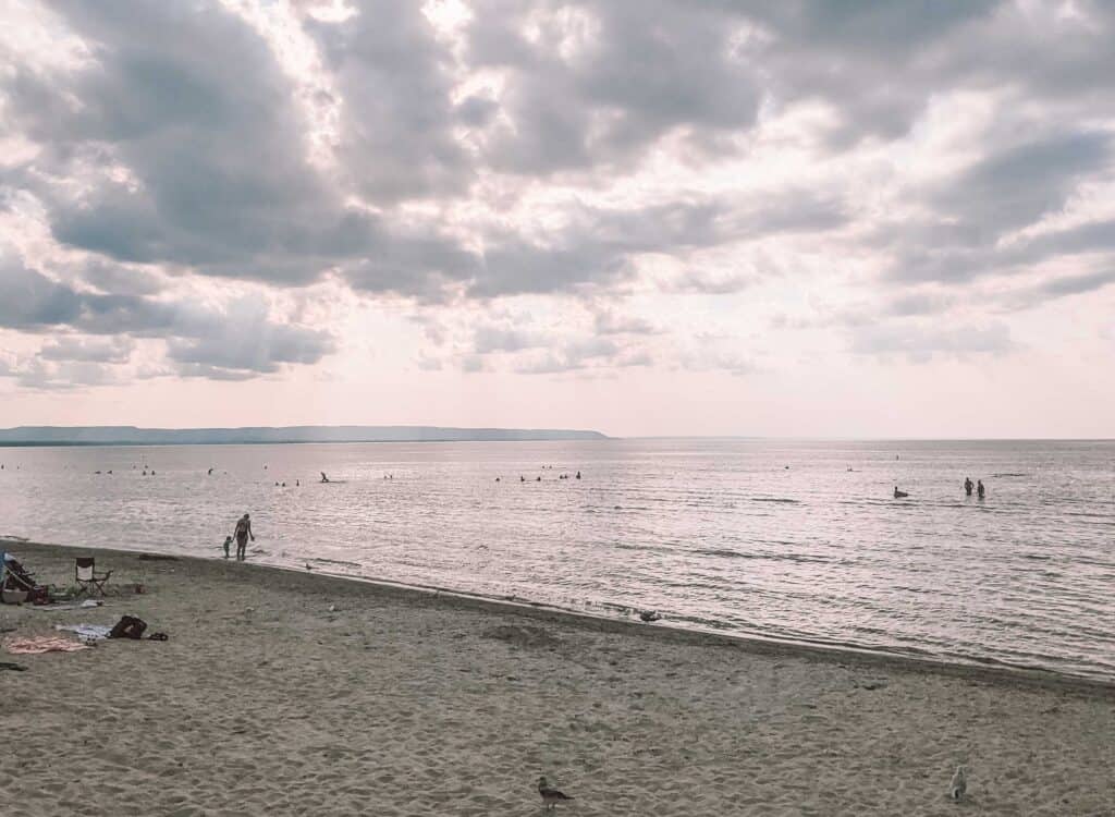 Sandy shoreline at Wasaga Beach Provincial Park along Georgian Bay. People swim and wade in the shallow water while dramatic clouds drift across the sky. The long freshwater beach stretches along the shoreline, with the Niagara Escarpment visible in the distance.