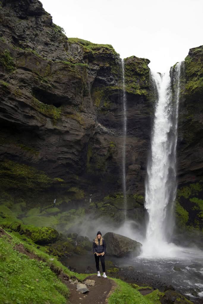 Visitor standing on a rocky path in front of Kvernufoss waterfall in southern Iceland, surrounded by steep, moss-covered cliffs and mist rising from the pool below.