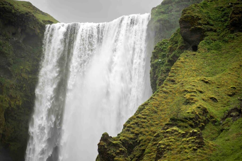 Close-up of Skógafoss waterfall in southern Iceland, with water plunging over a steep cliff beside a bright green, moss-covered hillside.