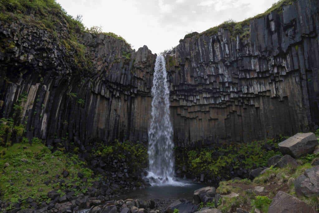Svartifoss waterfall cascading into a rocky pool in Skaftafell, Iceland, surrounded by tall, dark hexagonal basalt columns and patches of green vegetation.