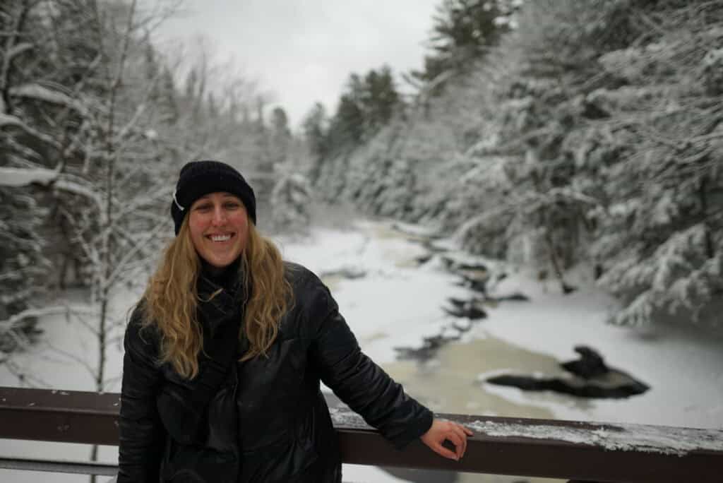 A smiling woman wearing a black winter coat and knit hat stands on a bridge overlooking a partially frozen creek surrounded by snow-covered trees in Arrowhead Provincial Park, Ontario.