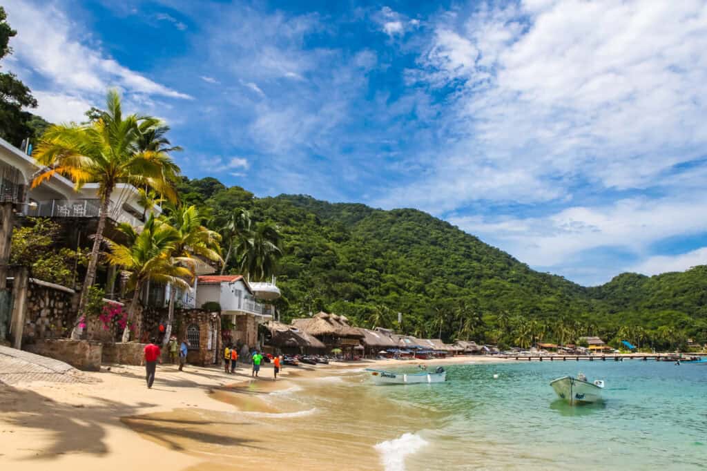 A sandy beach lined with palm trees and low buildings sits beside clear turquoise water, with small boats floating near shore and lush green hills rising behind the coastline under a bright, partly cloudy sky.