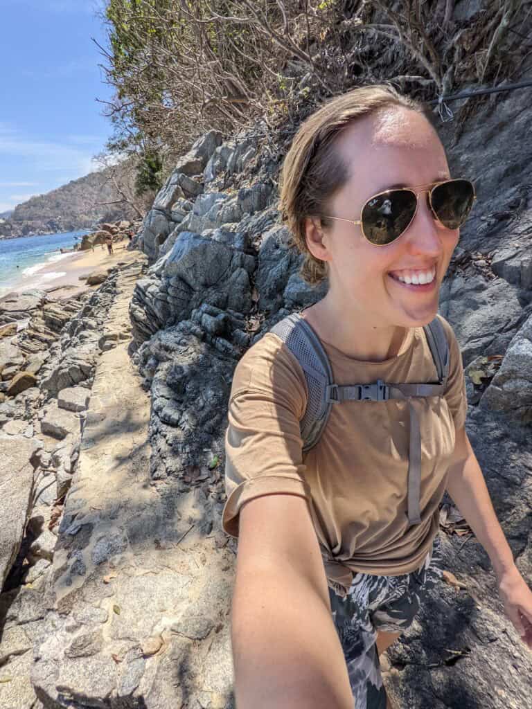 Person hiking along a narrow rocky coastal trail beside the ocean, wearing a backpack and sunglasses, with cliffs and beach visible below.