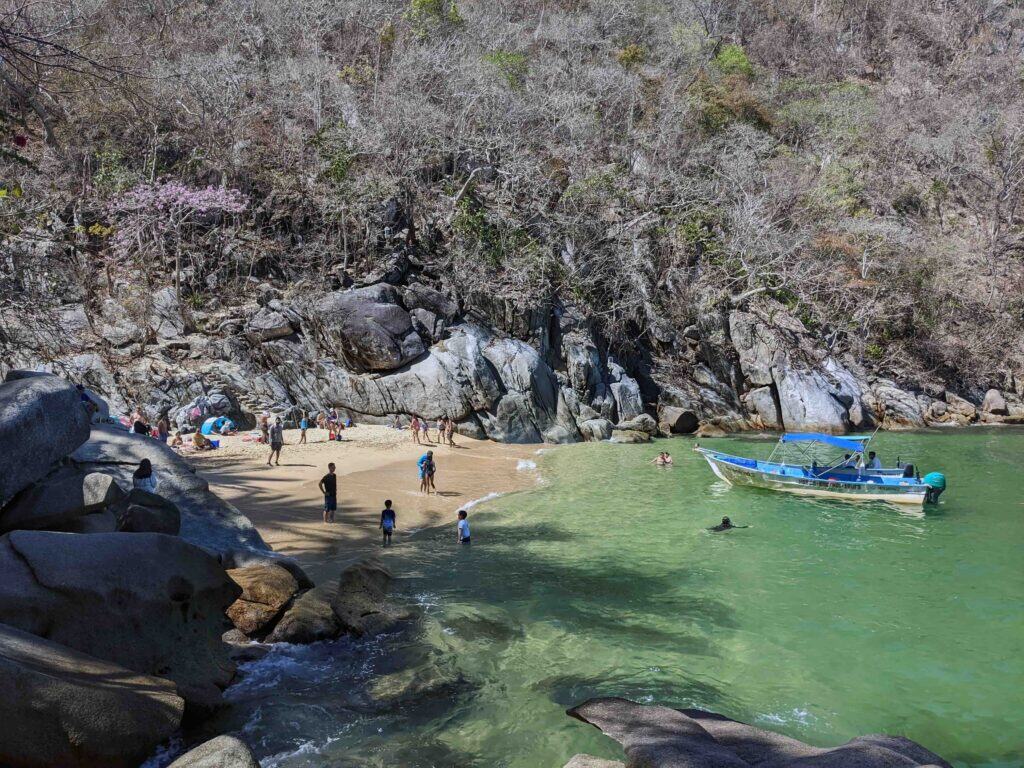 Small sandy cove with green water, people wading and swimming near shore, and a small motorboat anchored close to the beach, surrounded by rocky cliffs and dry coastal forest.