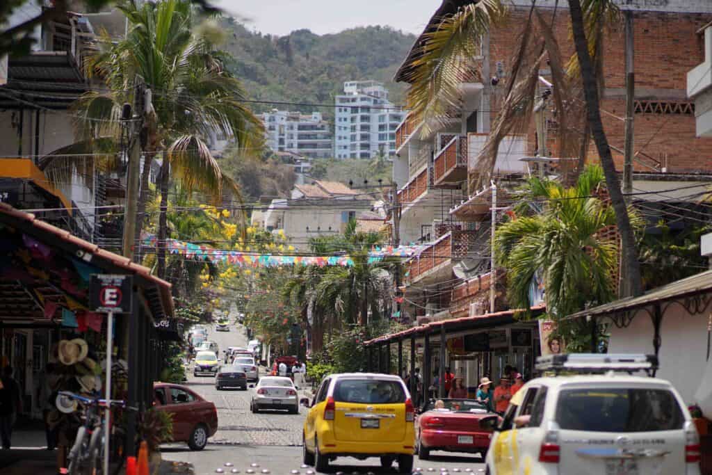 Narrow street in Puerto Vallarta’s Zona Romántica with cars, pedestrians, palm trees, colorful papel picado overhead, and hillside apartment buildings in the distance.