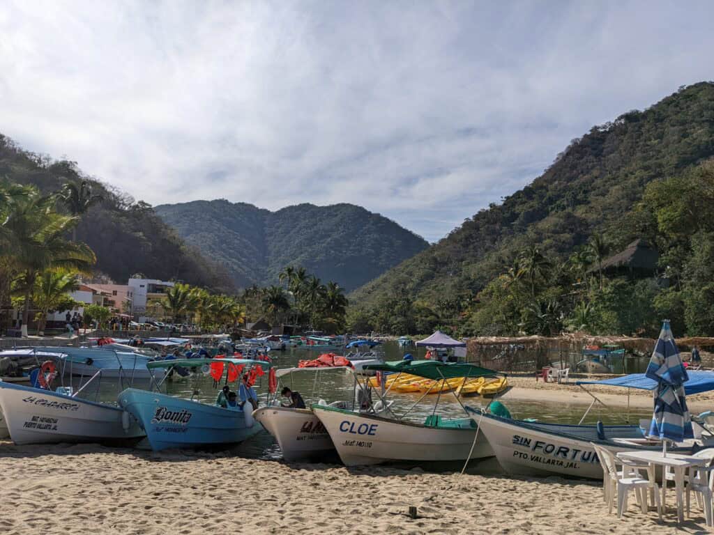 Small fishing boats are pulled up on a sandy beach in Puerto Vallarta, with calm water behind them, palm trees along the shore, and green hills rising in the background under a lightly clouded sky.