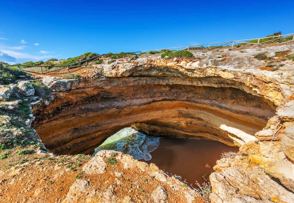 The famous Benagil Cave viewed from above, showing the natural skylight opening, golden limestone cliffs, and beach below in the Algarve, Portugal.
