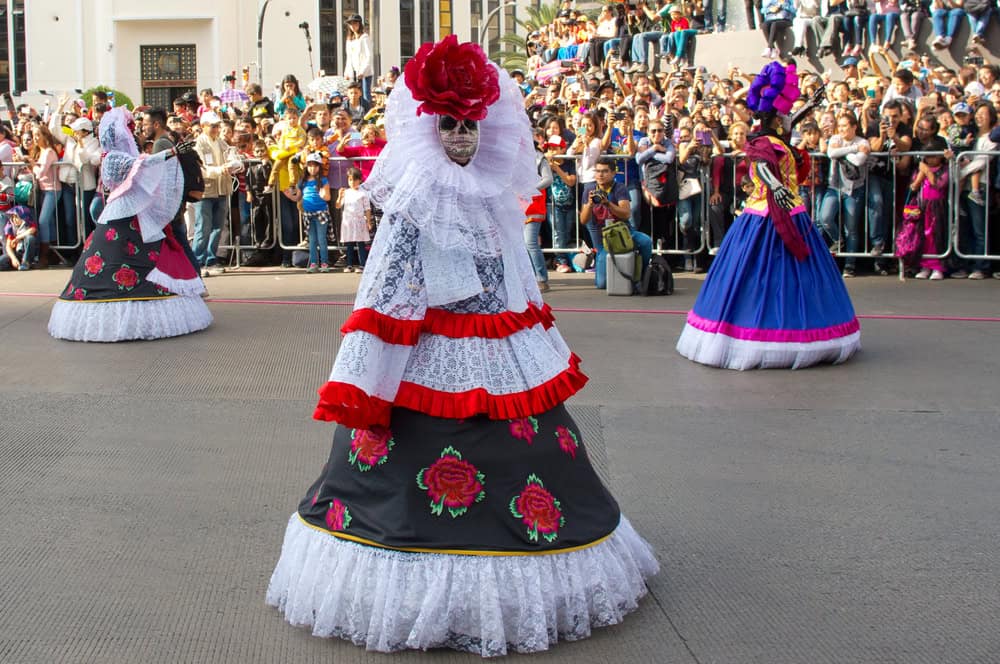women dressed in catrina costumes and walking past huge crowds during the day of the dead parade in mexico city.