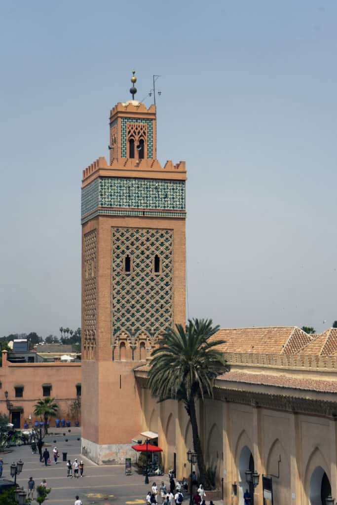 Koutoubia Mosque minaret in Marrakech, Morocco, featuring geometric brickwork and turquoise tile details, with palm trees and visitors in the square below.
