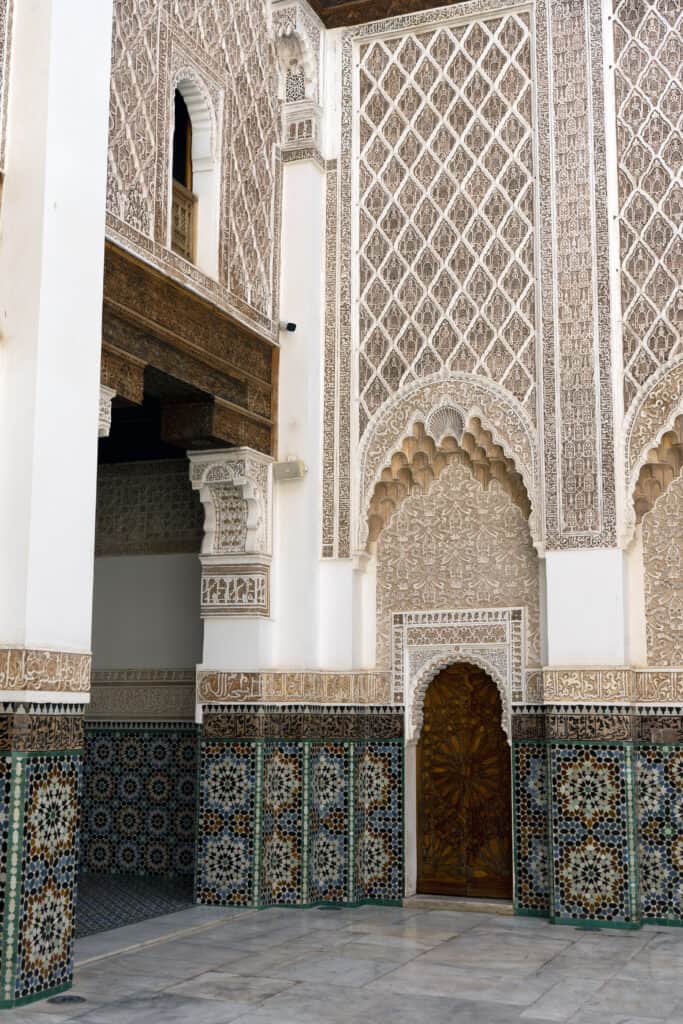Intricate carved plasterwork and colourful zellige mosaic tiles decorating the walls and arched doorway of a historic Moroccan palace.