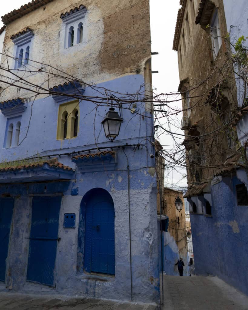 Narrow cobblestone street in Chefchaouen, Morocco, lined with blue-painted buildings, wooden doors, and traditional lanterns, with two pedestrians walking downhill.