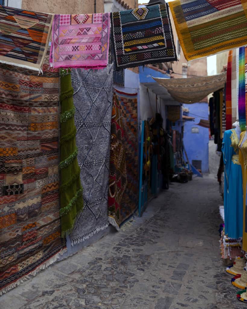 Colourful woven Moroccan rugs hanging outside shops along a narrow cobblestone street in Chefchaouen, Morocco, with blue-painted buildings in the background.