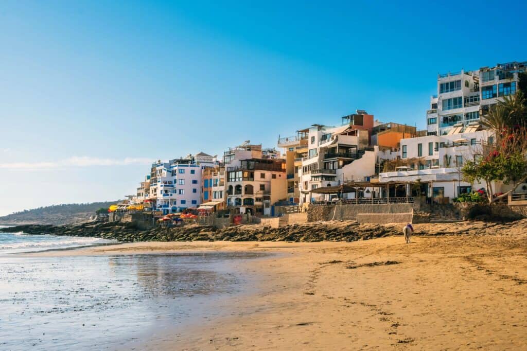 Sandy beach in Taghazout, Morocco, with Atlantic Ocean waves along the shoreline and white apartment buildings, hotels, and restaurants overlooking the water under a clear blue sky.