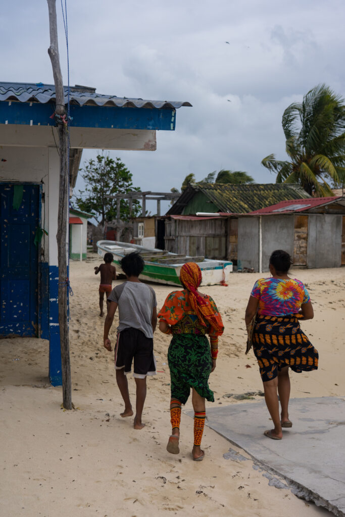 Three people walking along a sandy street in a coastal village, passing colourful houses, wooden buildings, fishing boats, and palm trees under a cloudy sky.