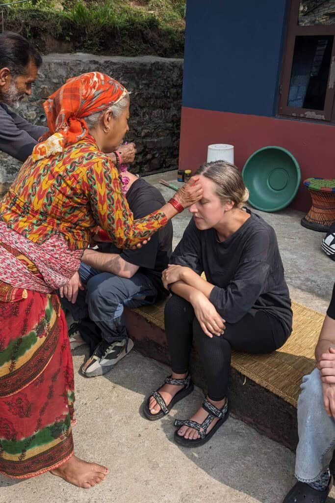 Woman wearing colourful traditional clothing and an orange headscarf placing a tika on a seated traveller's forehead during a welcome ceremony at a homestay in Barang, Nepal.