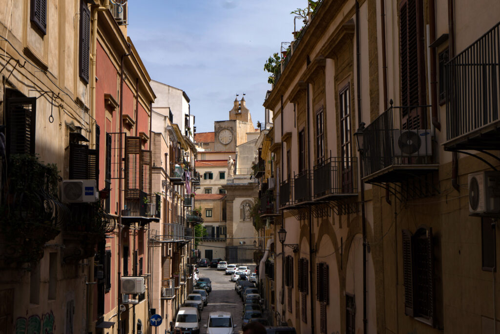 a street in Palermo lined with cars and colourful buildings that are pastel pink and yellow, with balconies overlooking the street