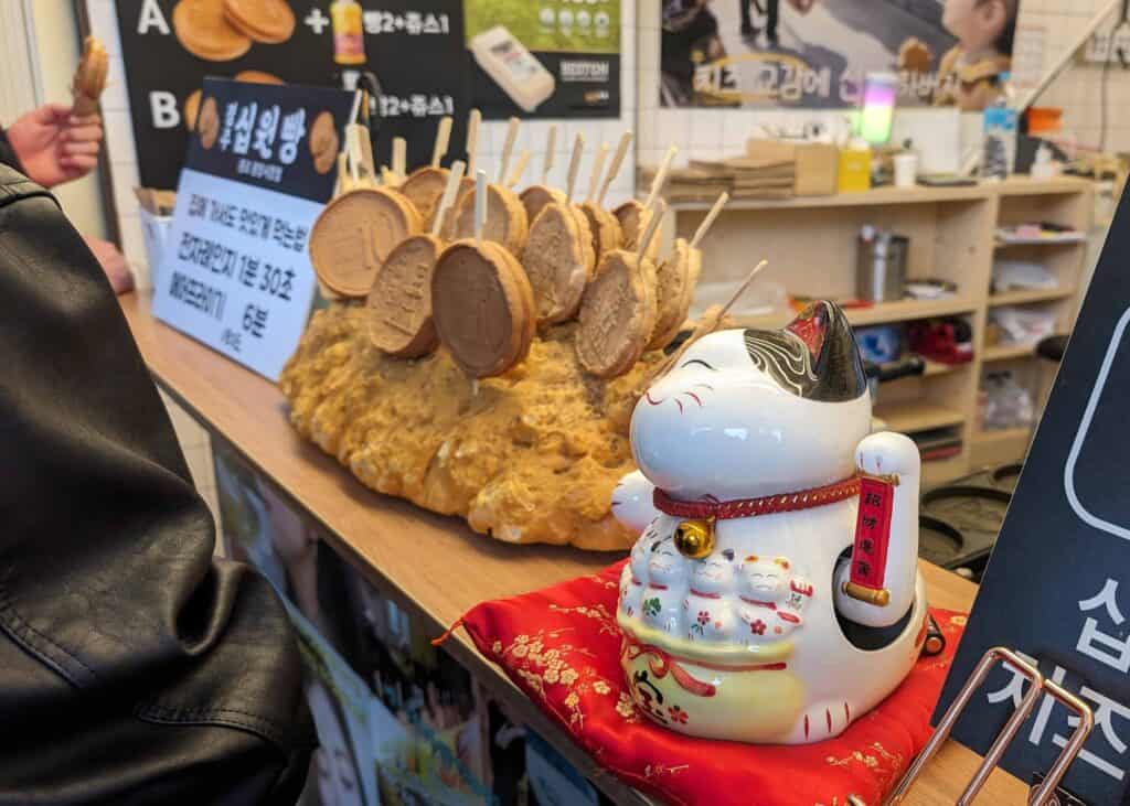 Counter at a food stall in Gwangjang Market with fish-shaped pastries on sticks and a ceramic lucky cat figurine displayed in the foreground.