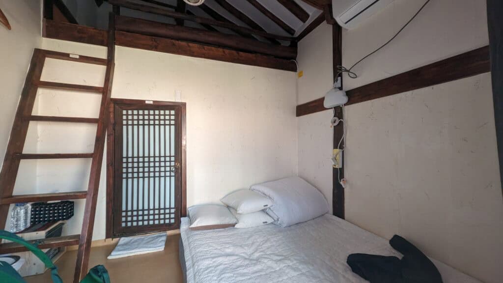 a small hanok room with. a large sleeping mat, white walls, and wooden accents, as well as a ladder leading up to a cozy attic