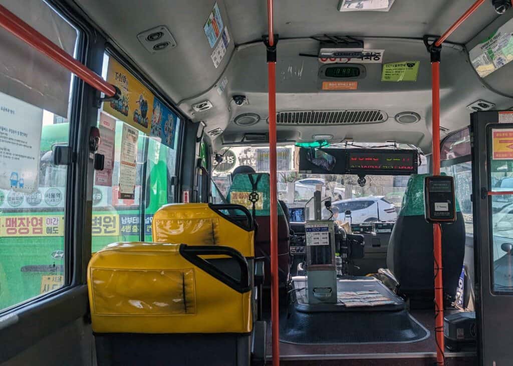 Interior of a public bus in Seoul, with yellow seats, handrails, and the driver’s area visible at the front.