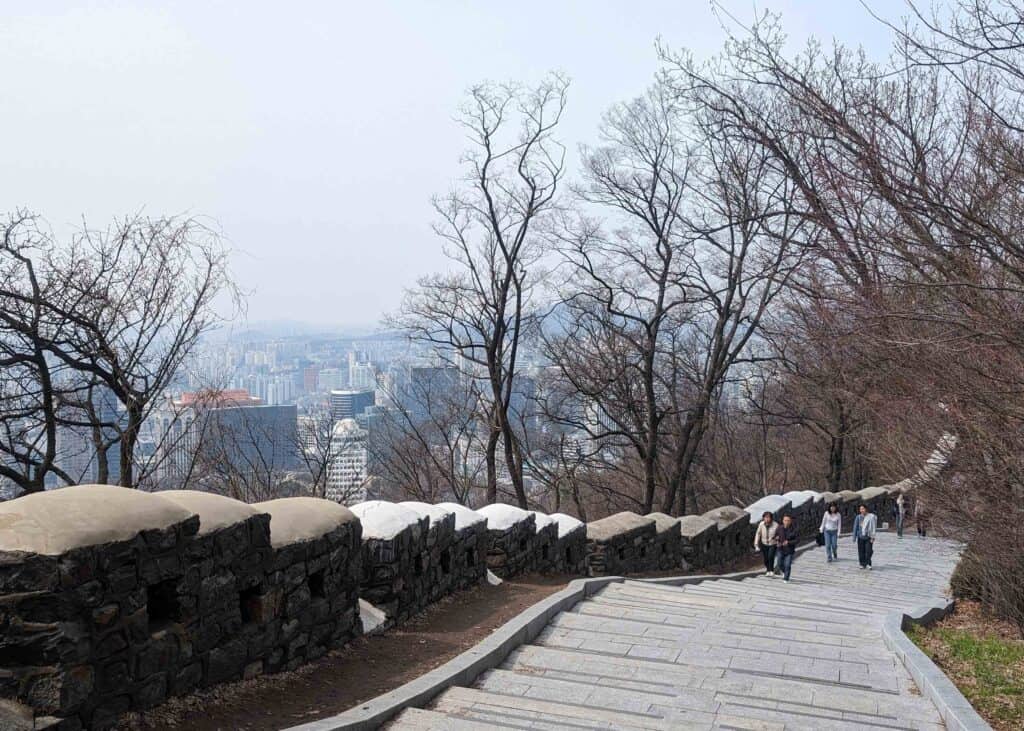 Stone path along the Seoul City Wall on Namsan Mountain, with people walking uphill and views of the Seoul skyline through bare trees.