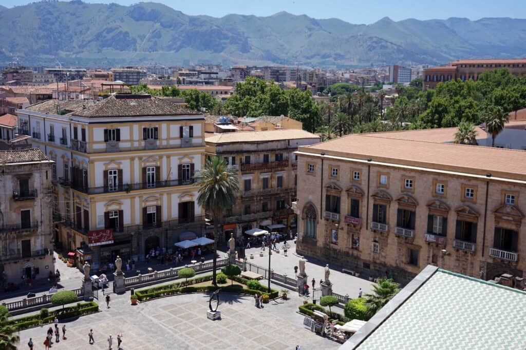 View over a square in Palermo with historic buildings, palm trees, and Mount Pellegrino rising in the background.
