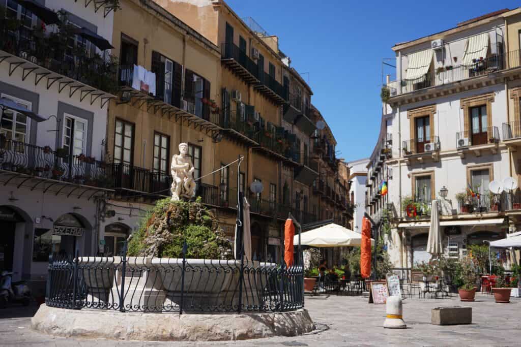 Fountain with a marble statue in Piazza San Domenico in Palermo, surrounded by historic apartment buildings, balconies, and outdoor cafés in the Kalsa district.