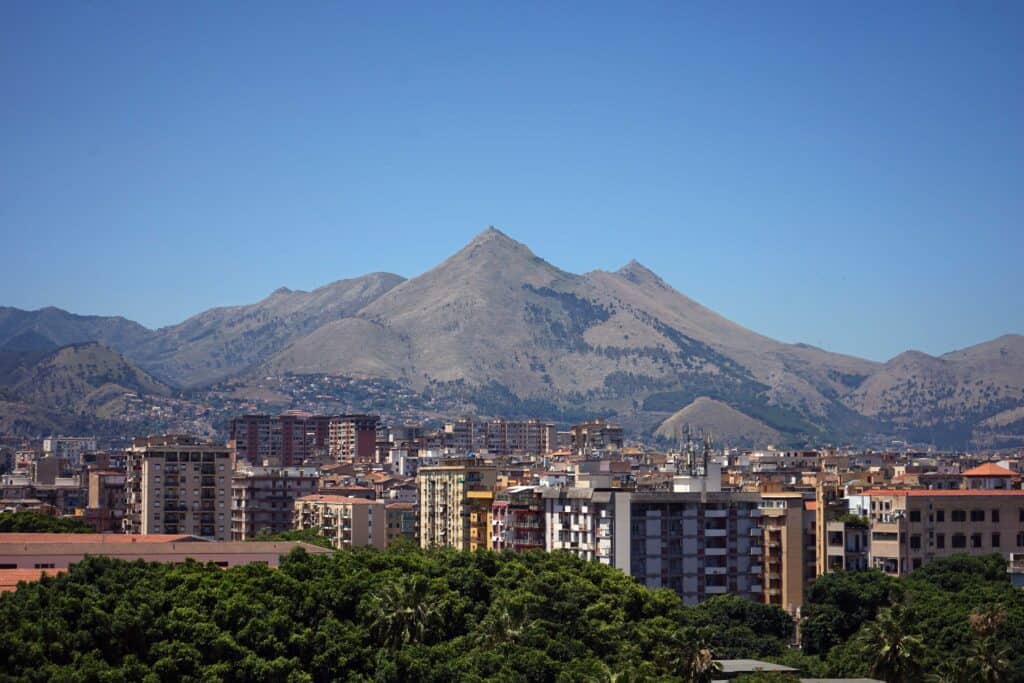 View over Palermo’s cityscape with residential buildings in the foreground and the mountains surrounding the city rising in the background under a clear blue sky.