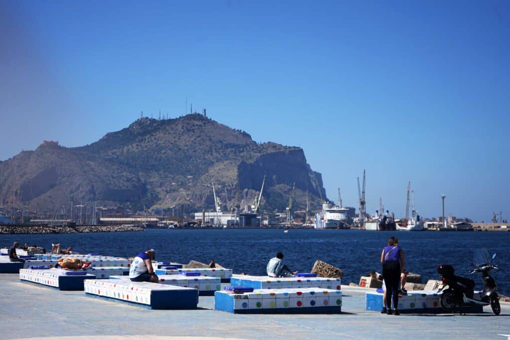 People relaxing on benches along Palermo’s waterfront with the harbor, ships, and Mount Pellegrino in the background.