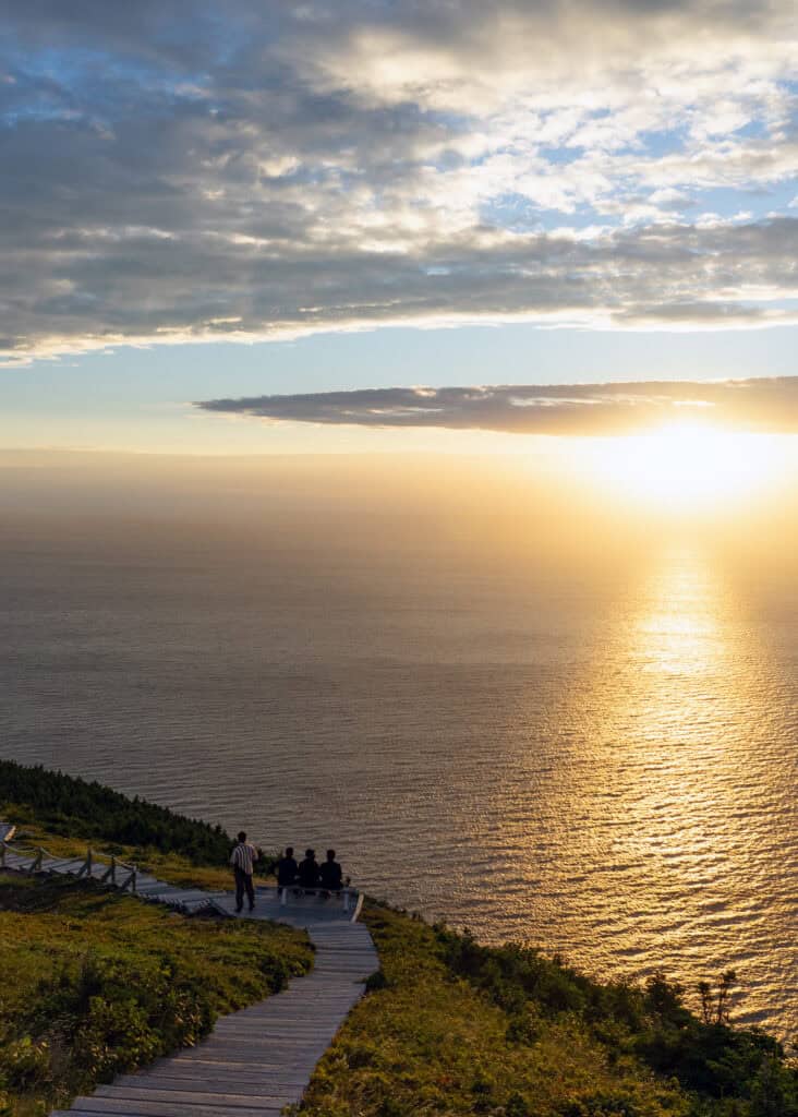 people sitting on a boardwalk looking out over the ocean at sunset time on cape breton