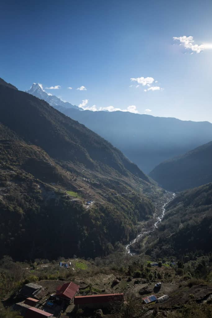 View of a Himalayan valley during the Poon Hill Trek in Nepal, with a river winding through the valley below and snow-capped mountains rising in the distance beneath a clear blue sky.
