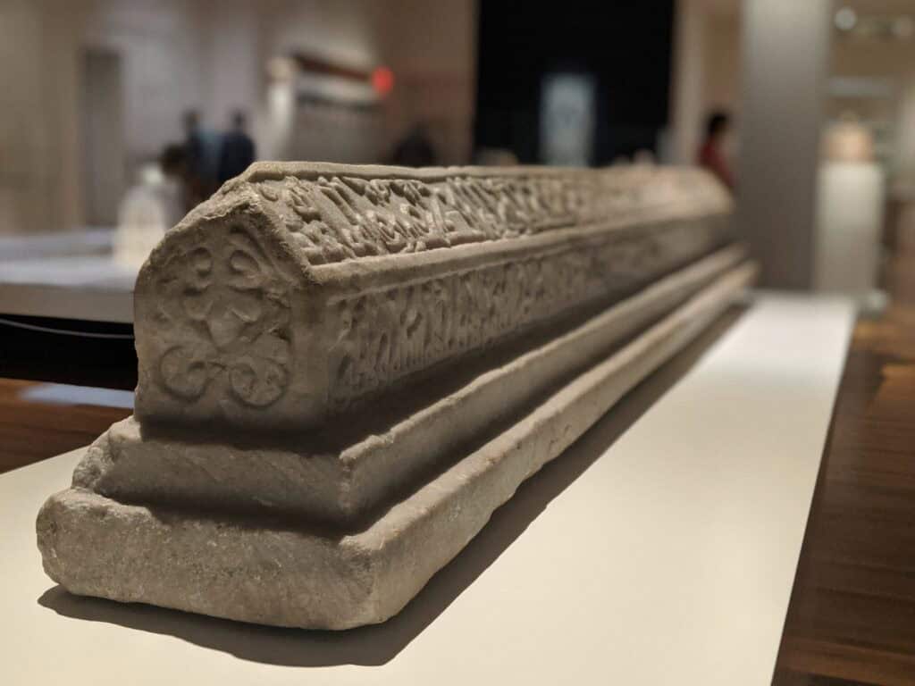 Ancient carved stone sarcophagus displayed inside the Aga Khan Museum in Toronto. The rectangular artifact features intricate decorative patterns and inscriptions carved into its surface, with the museum gallery softly blurred in the background.