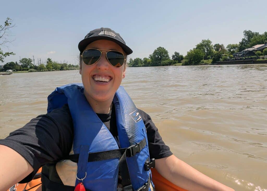 a woman smiling into the camera while floating in an orange canoe on a muddy river, paddling in windsor-essex