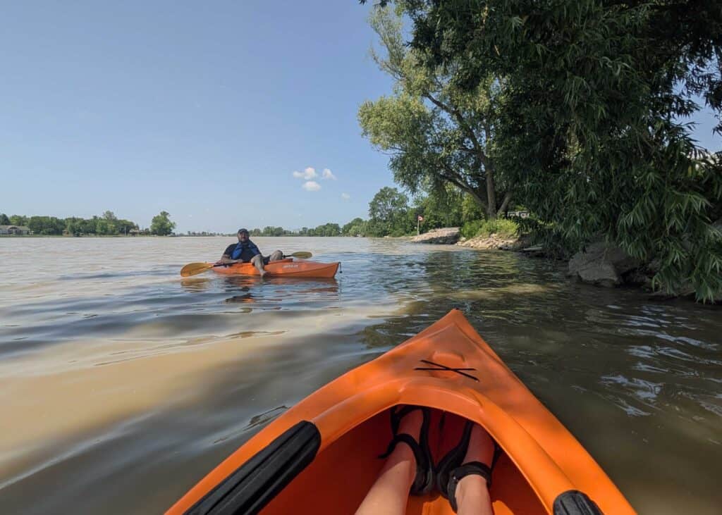the front of orange kayak floating on a river next to trees overhanging, and a man sitting in a kayak floating nearby