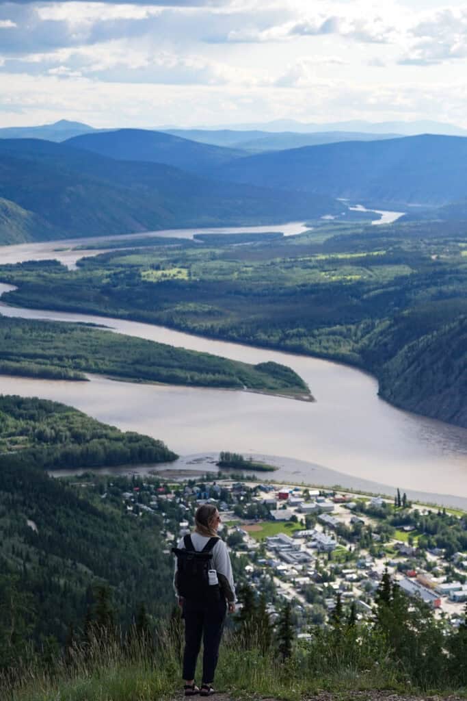 a woman standing on the edge of a lookout point with views of rolling hills and dawson city
