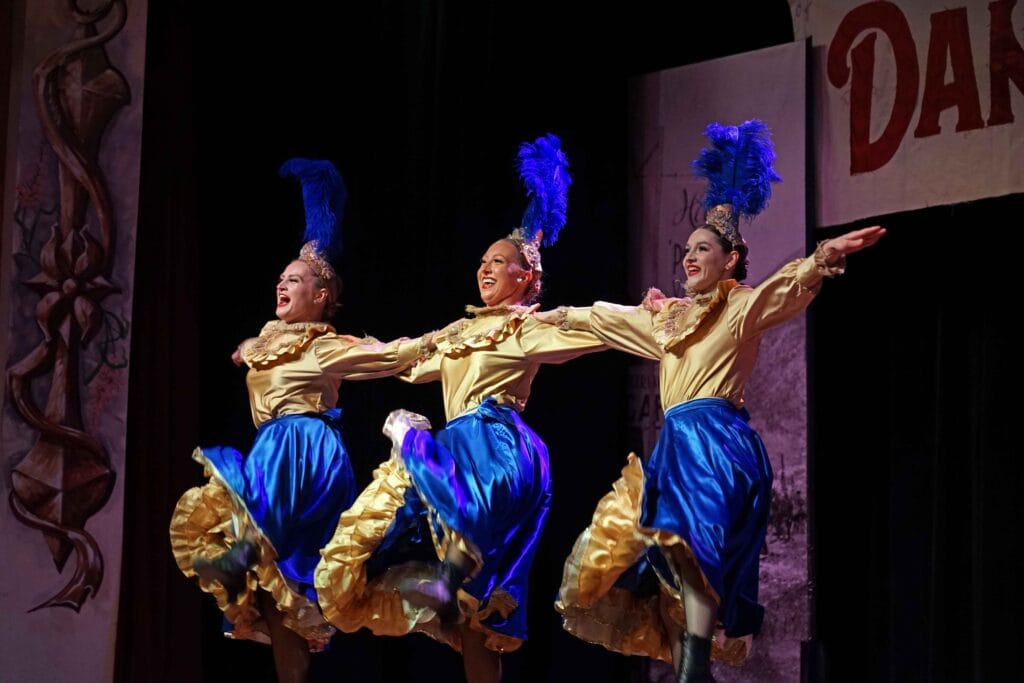 three can can dancers standing in a row, with their arms extended, wearing blue skirts and gold tops, with feather hats