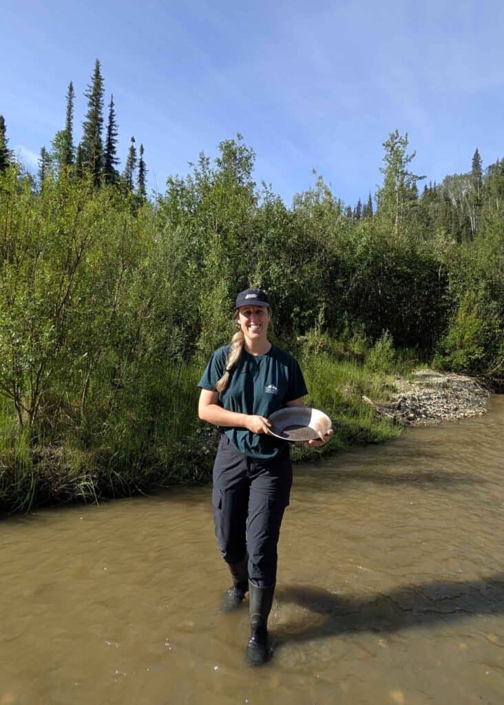 a woman wearing a cap and holding a gold pan, wading across a small stream toward the camera