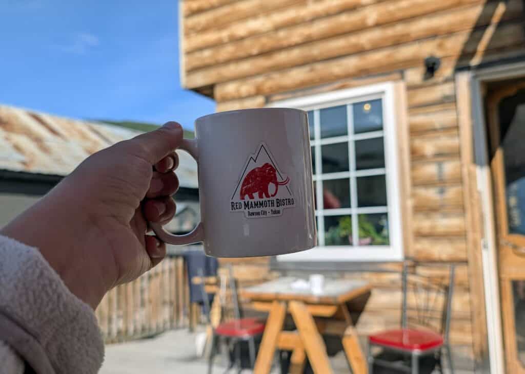 a hand holding a mug that reads "red mammoth bistro" in front of an old log building