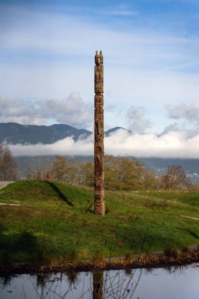A tall totem pole on a lawn next to a pond, with mountains and fog in the distance, on the UBC campus.