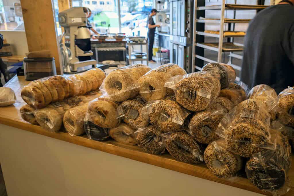 Stacks of freshly baked Montreal-style bagels wrapped in clear bags on a wooden counter inside Bullet Hole Bagels, with bakers working in the background.