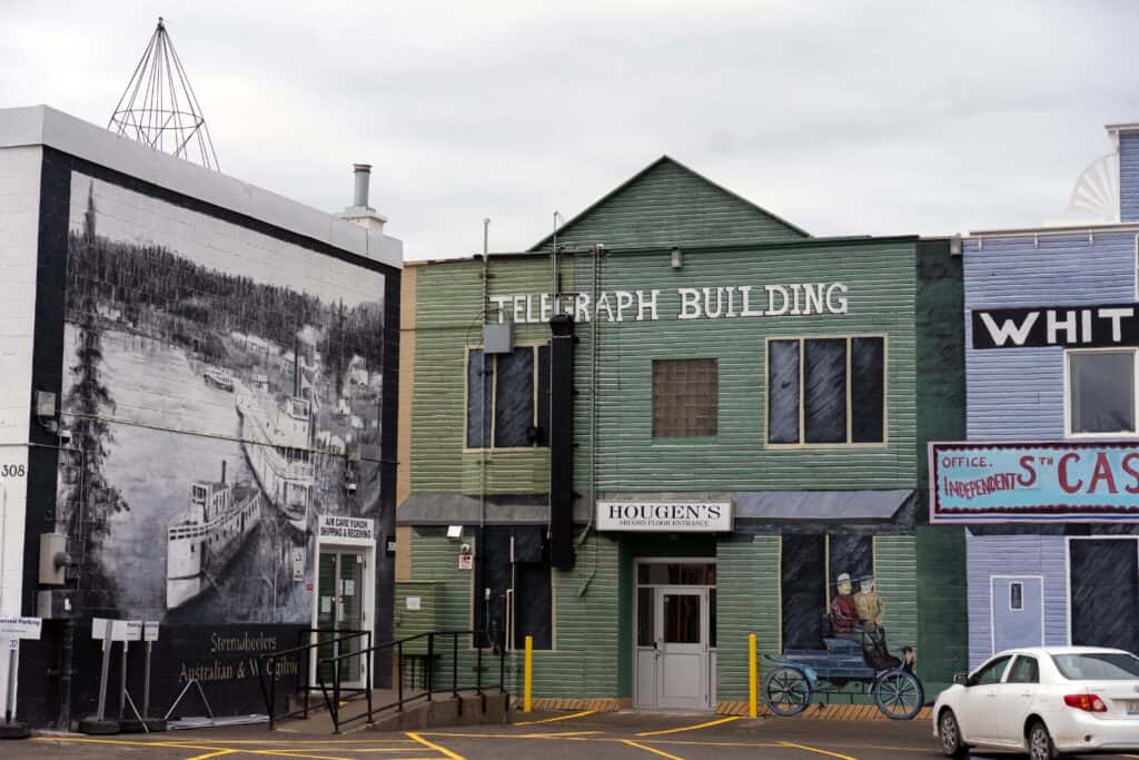 Historic Telegraph Building in downtown Whitehorse with green wooden siding, painted murals depicting early river travel, and neighbouring heritage façades on an overcast day.