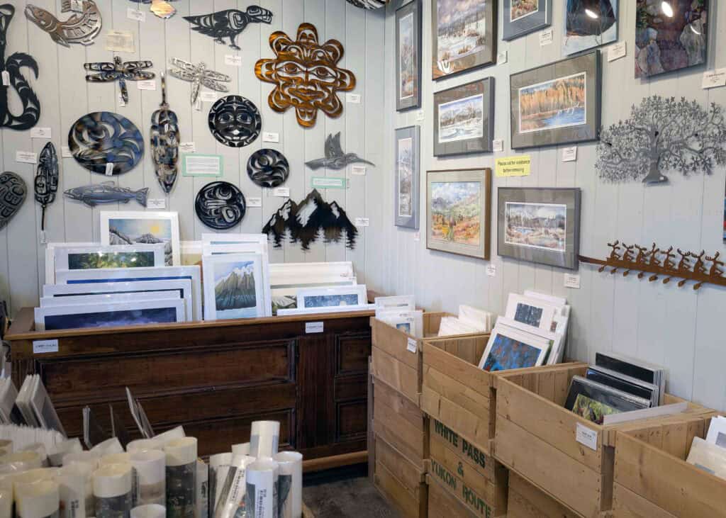 Interior of North End Gallery in Whitehorse, showing walls filled with framed landscape art and metal wall sculptures, with wooden bins and shelves displaying prints and artwork by Yukon and northern artists.